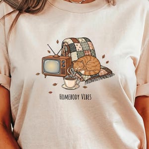 May include: Beige t-shirt featuring a cozy illustration. A vintage TV, a patchwork blanket, and a sleeping orange tabby cat are depicted. A cup of coffee steams beside the text "HOMEBODY VIBES". Autumn leaves are scattered around the scene.