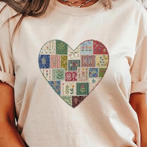 May include: A light beige t-shirt featuring a large heart design. The heart is composed of various colorful patchwork squares with floral and geometric patterns. The shirt has short sleeves and a crew neck.