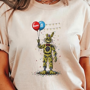 May include: Beige t-shirt featuring a yellow character holding red and blue balloons with the text "Name 8". The character is surrounded by confetti and string lights. The design is a cartoon style.