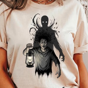 May include: Ivory-colored t-shirt featuring a black and gray illustration of a person holding a lantern, with a shadowy figure looming behind. The illustration has a horror theme, with the word "Ivory" printed at the bottom.