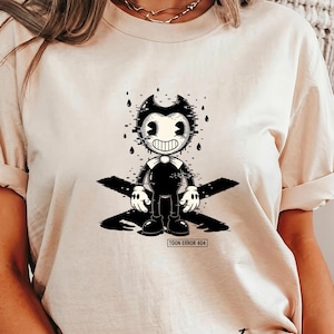 May include: Ivory-colored t-shirt featuring a black and white cartoon character with a glitch effect. The character has a wide smile and is standing on a black cross-shaped design. The text "TOON ERROR 404" is below the character.