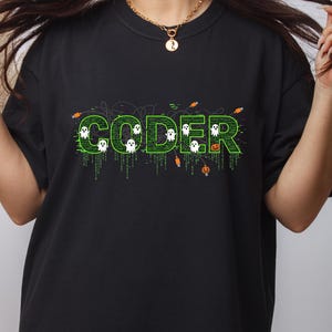 May include: Black t-shirt with the word "CODER" in green, featuring a Halloween-themed design. The word is filled with binary code and small ghost graphics. Orange jack-o'-lanterns and cable graphics are also included in the design.