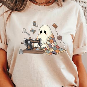 May include: Ivory-colored t-shirt featuring a whimsical illustration of a ghost sewing a quilt on a vintage sewing machine. The design includes spools of thread, scissors, and a thimble. The word "Ivory" is printed at the bottom.