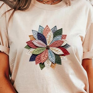 May include: Beige t-shirt featuring a colorful, layered floral design. The flower has multiple petals in shades of red, pink, blue, green, and gold. The shirt has short sleeves and a crew neck.