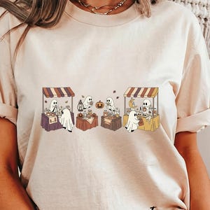 May include: Ivory-colored t-shirt featuring a whimsical Halloween market scene. The design showcases cartoon ghosts at market stalls with pumpkins, candles, and autumn leaves. The word "Ivory" is printed in a script font at the bottom right.