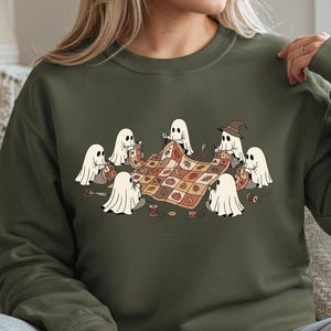 May include: Military green sweatshirt featuring a whimsical illustration of several ghost figures gathered around a quilt. The ghosts are sewing and crafting, with scissors, spools of thread, and a witch's hat. The quilt has a fall-themed pattern.