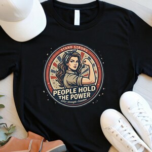 May include: Black t-shirt featuring a circular graphic with a woman flexing her arm, the text "PEOPLE HOLD THE POWER", and the words "unity, strength, freedom". A white baseball cap, sunglasses, and sneakers are also visible.