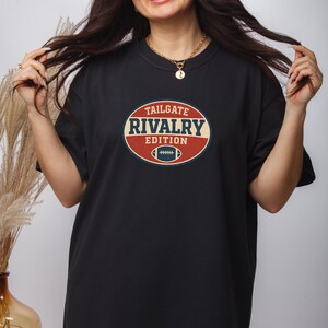 May include: Black t-shirt featuring a retro-style oval design with the text "TAILGATE RIVALRY EDITION" and a football graphic. The design is in red, blue, and cream colors. A gold necklace is visible.