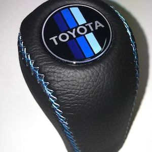 May include: Black leather shift knob with blue stitching. The top features a circular emblem with the Toyota logo and blue stripes. The shift knob is designed for a vehicle's gear shift.