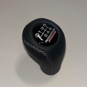 Op de afbeelding: Een zwarte lederen versnellingspookknop met een handgeschakeld patroon van zes versnellingen inclusief achteruit, witte stiksels aan de zijkant en een ronde inzet aan de bovenzijde met de tekst MAZDASPEED MOTORSPORTS in witte en rode letters.