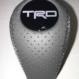 Op de afbeelding: Grijze lederen versnellingspookknop met de witte letters "TRD" op een zwarte cirkelvormige achtergrond. De knop heeft een geperforeerde textuur en zilveren stiksels. Auto-accessoire.