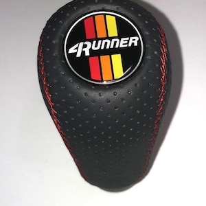 May include: Black shift knob with red stitching. The top features a circular emblem with the text "4Runner" and a design of red, orange, and yellow stripes. The knob has a perforated texture.