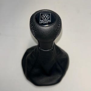 Könnte beinhalten: Ein schwarzer Schaltknauf aus Leder mit strukturierter Oberfläche und einem runden Einsatz auf der Oberseite, der das Volkswagen-Logo und den Schriftzug MOTOR SPORT in Weiß zeigt. Der Knauf ist mit einer schwarzen Ledermanschette mit sichtbaren Nähten verbunden.