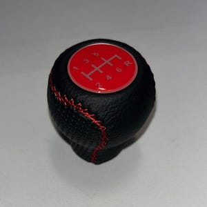 Peut inclure: Pommeau de levier de vitesses noir avec surpiqûres rouges et un dessus rouge. Le dessus comporte un schéma de changement de vitesse blanc avec les chiffres 1, 2, 3, 4, 5, 6 et R.