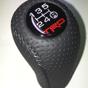May include: Black leather gear stick knob with a perforated surface and white stitching. The top has a black emblem with white gear shift numbers and the red "TRD" logo.
