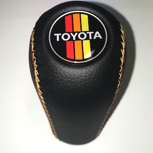 May include: Black shift knob with the Toyota logo in a circular emblem. The emblem features red, orange, and yellow stripes. The knob has yellow and orange stitching.