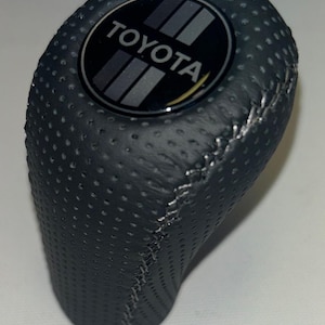 May include: A dark gray, perforated leather shift knob with silver stitching. The top features a black emblem with the TOYOTA logo and three gray stripes. The knob has a rounded, ergonomic shape.