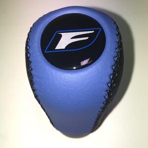 Könnte beinhalten: Blauer und schwarzer Schalthebelknauf mit gestepptem Lederdesign. Der Knauf hat ein schwarzes Emblem mit einem weißen "F"-Logo, das blau umrandet ist.