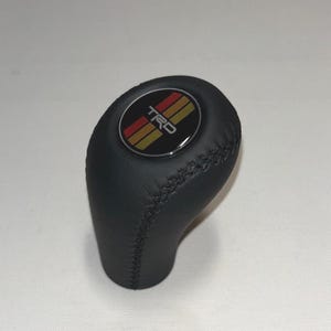 May include: A black leather gear shift knob featuring a circular emblem on top. The emblem displays the text TRD in white letters over three vertical stripes in red, orange, and yellow. The knob has visible black stitching along the side seams.