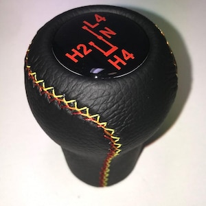 Könnte beinhalten: Schwarzer Schalthebelknauf mit roten und gelben Nähten. Die Oberseite des Knaufs ist schwarz mit roten Buchstaben, die die Gangpositionen angeben: H2, L4, N und H4.