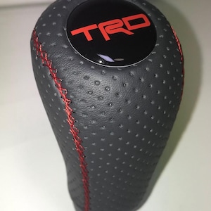 Peut inclure: Un pommeau de levier de vitesses en cuir noir perforé avec des coutures rouges. Le dessus présente un cercle noir avec les lettres rouges "TRD". Le pommeau est conçu pour le changement de vitesse d'un véhicule.