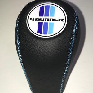 Könnte beinhalten: Schwarzer Schalthebelknauf mit blauen Nähten. Die Oberseite hat einen weißen Kreis mit dem Text "4RUNNER" und blauen vertikalen Streifen. Der Knauf hat eine strukturierte Oberfläche.
