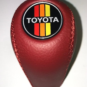 May include: A red leather shift knob with a stitched seam. The knob features a circular emblem with the TOYOTA logo and vertical stripes in red, orange, and yellow.