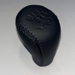 May include: Black leather shift knob with embossed gear pattern and "TRD" logo. The knob has a stitched seam and a rounded shape, designed for a vehicle's gear shift lever.