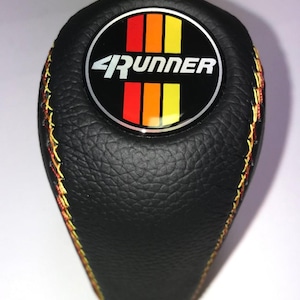 May include: Black leather shift knob with the word "4Runner" in white lettering on a circular emblem. The emblem features vertical stripes in red, orange, and yellow. The knob has yellow and red stitching.