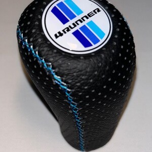 May include: Black shift knob with a textured surface and blue stitching. The knob features a white circular emblem with the text "4RUNNER" and blue stripes. Automotive accessory.