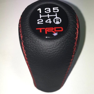 May include: Black gear stick knob with red stitching. The top has a black circle with white gear shift numbers and the red TRD logo. The shift pattern is 1, 3, 5, 2, 4, and R.
