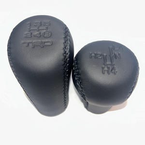 May include: Two black leather gear shift knobs. One is elongated with the numbers 1, 3, 5, 2, 4, R, and the letters TRD embossed on it. The other is round with the letters H2, L, N, H4 embossed on it.