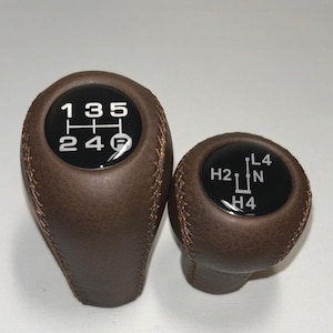Könnte beinhalten: Zwei braune Lederschaltknäufe mit sichtbaren Nähten. Der größere Schaltknauf hat eine schwarze Oberseite mit einem weißen 1-3-5 und 2-4-R Schaltschema. Der kleinere hat eine schwarze Oberseite mit einem weißen H2-N-L4 und H4 Schema.