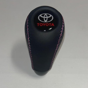 May include: A black gear shift knob featuring a smooth top surface with the white Toyota logo and red Toyota text. The sides of the knob are textured and accented with pink stitching that runs vertically along the seams of the black material.