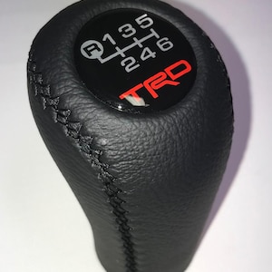 May include: Black leather gear shift knob with a stitched seam. The top features a black circle with a white gear shift pattern and the red letters "TRD".