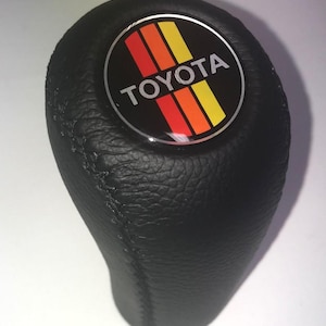 May include: Black leather Toyota gear stick knob with a circular emblem. The emblem features the Toyota logo in white text, with red, orange, and yellow stripes. The gear stick knob is designed for a vehicle's gear shift.