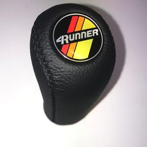 Peut inclure: Pommeau de levier de vitesses noir avec une texture en cuir cousu. Le bouton présente un emblème circulaire avec le mot "4Runner" en blanc et des rayures rouges, orange et jaunes. L'emblème est sur fond noir.
