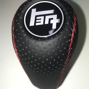 May include: Black shift knob with red stitching. The knob has a perforated texture and a circular emblem with white text on a black background. The emblem features a stylized logo.