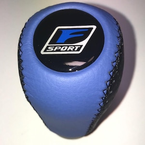 May include: Blue and black car gear shift knob with the "F SPORT" logo in white and blue on a black background. The knob is made of leather and has black stitching.