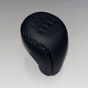 May include: Black leather-wrapped shift knob with a six-speed gear pattern. The knob has a stitched seam detail and a rounded, ergonomic shape. The gear pattern is embossed on the top surface.