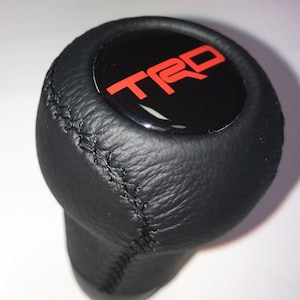 May include: Black leather shift knob with the red TRD logo on a black circular emblem. The shift knob has a stitched seam and a rounded shape. The background is a solid white.