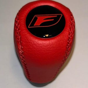 May include: A red leather shift knob with a black circular emblem featuring a red "F" logo. The shift knob has a stitched design along the sides and a dark gray base. Automotive accessory.