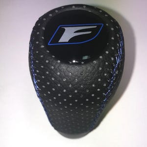May include: Black leather shift knob with a perforated texture and blue stitching. The top features a black circle with a silver and blue "F" logo. Automotive accessory.