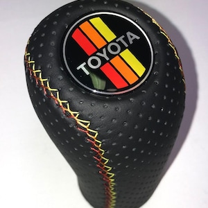 May include: Black perforated car gear shift knob with a stitched seam in yellow and red thread. The top features a circular emblem with the Toyota logo and stripes in red, orange, and yellow.