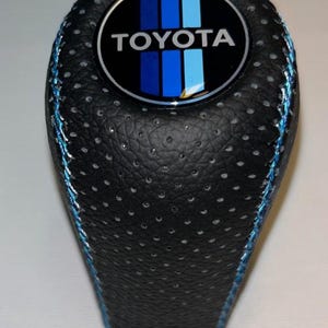 Peut inclure: Pommeau de levier de vitesse en cuir noir avec surpiqûres bleues. Le dessus présente un emblème circulaire avec le logo Toyota et des rayures bleues. Le pommeau a une surface texturée avec de petites perforations.