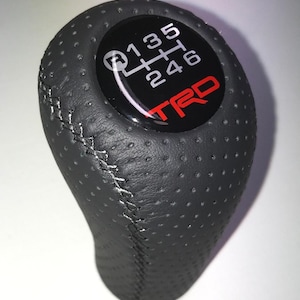Op de afbeelding: Zwarte, geperforeerde lederen versnellingspookknop met witte stiksels. De bovenkant heeft een zwarte wijzerplaat met een wit schakelpatroon en een rood "TRD"-logo. Een auto-accessoire.