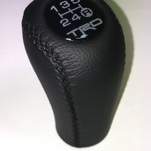 May include: Black leather gear stick knob with a white gear shift pattern and the letters "TRD". The knob has a stitched seam and a glossy black top.