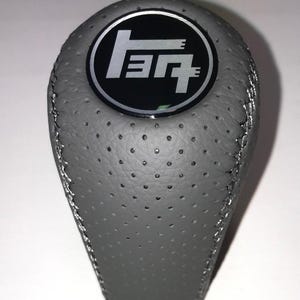 May include: Gray shift knob with a perforated surface and silver stitching. The top features a black circle with a white logo. The shift knob is designed for a vehicle's gear shift.