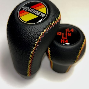 May include: Two black leather gear shift knobs with colorful stitching. The larger knob has a circular emblem with the word "4Runner" and a striped design. The smaller knob displays gear shift positions.