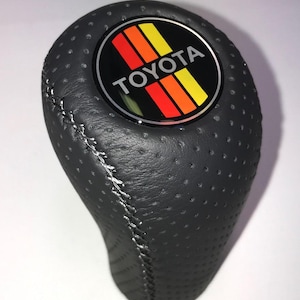 May include: A black, perforated leather shift knob with white stitching. The knob features a circular emblem with the Toyota logo and orange, yellow, and red stripes. The shift knob is designed for a vehicle's gear selector.
